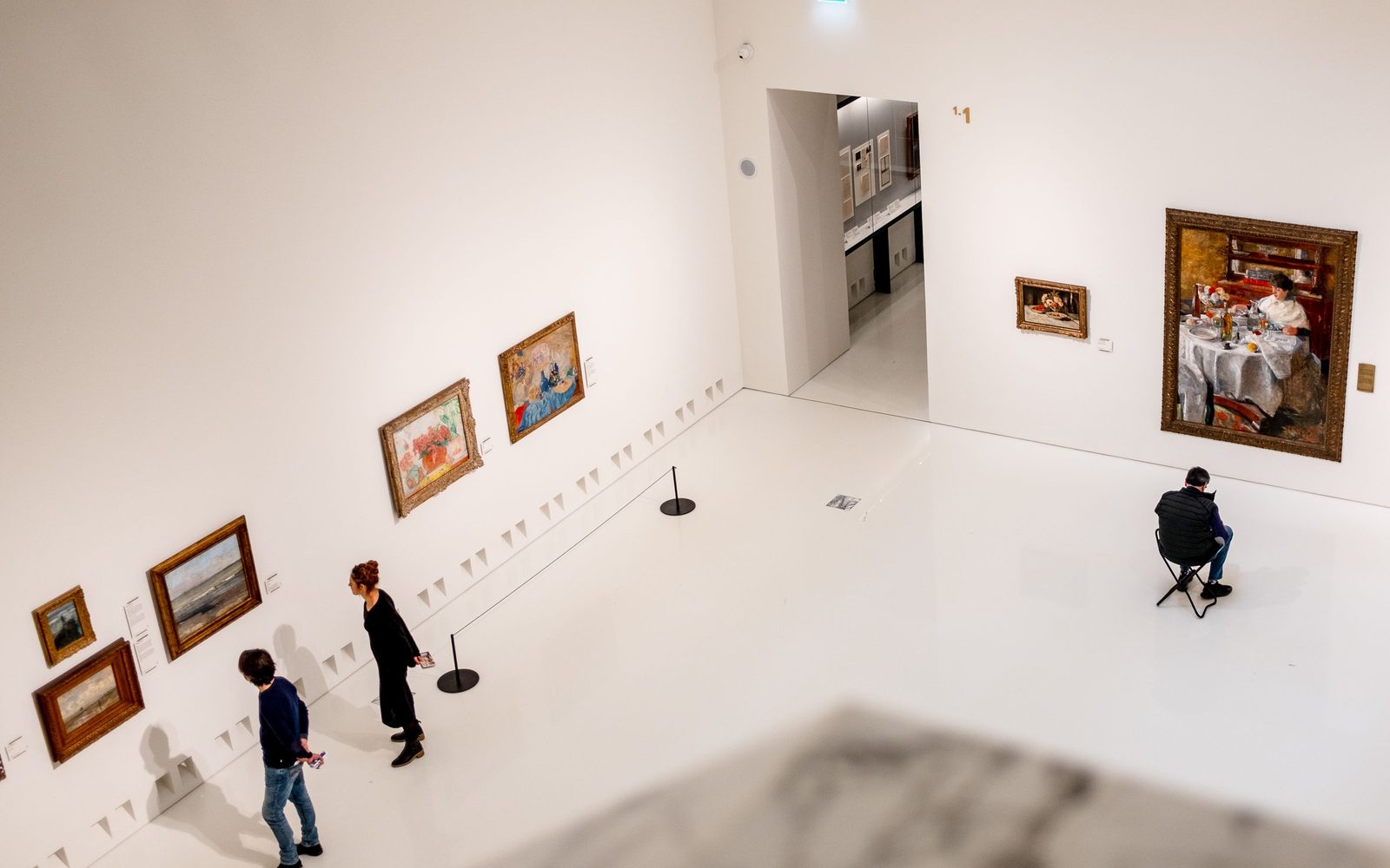 Des visiteurs regardent les tableaux d’une galerie aux murs blancs, près d’une porte qui ouvre sur une autre salle.